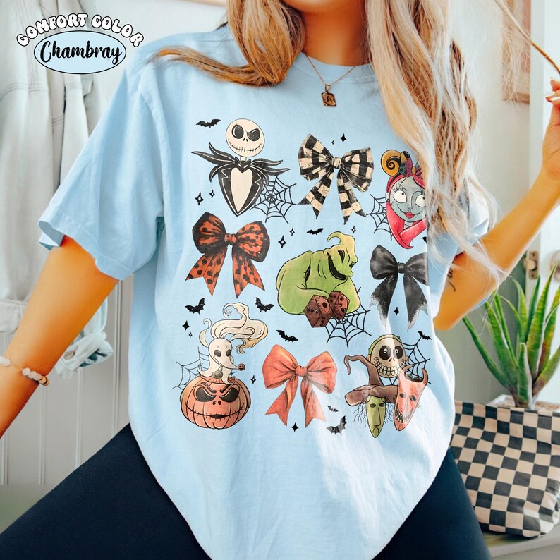 May include: A light blue t-shirt featuring a graphic print of characters and bows from The Nightmare Before Christmas, including Jack Skellington, Sally, Oogie Boogie, Zero, and various spider webs, with the text Comfort Color Chambray.