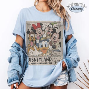 Puede incluir: Una camiseta azul claro con un estampado gr&aacute;fico rectangular de personajes de Disney que incluye a Mickey Mouse, Minnie Mouse, el Pato Donald, Goofy y Pluto. El texto dice Disneyland Where Dreams Come True 2024. Se ve una etiqueta Comfort Color Chambray.