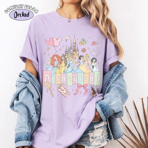 Puede incluir: Una camiseta lila Comfort Colors con un gr&aacute;fico de Disneyland. El dise&ntilde;o incluye princesas de Disney, un castillo y la palabra "Disneyland" en colores pastel. Gr&aacute;ficos adicionales incluyen palomitas de ma&iacute;z, un lazo y una varita m&aacute;gica.