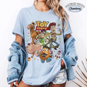 May include: Light blue Comfort Colors t-shirt featuring a graphic of characters from the animated film "Toy Story." The design includes Woody, Buzz Lightyear, Mr. Potato Head, and other characters. The words "Toy Story" are printed in yellow and red.
