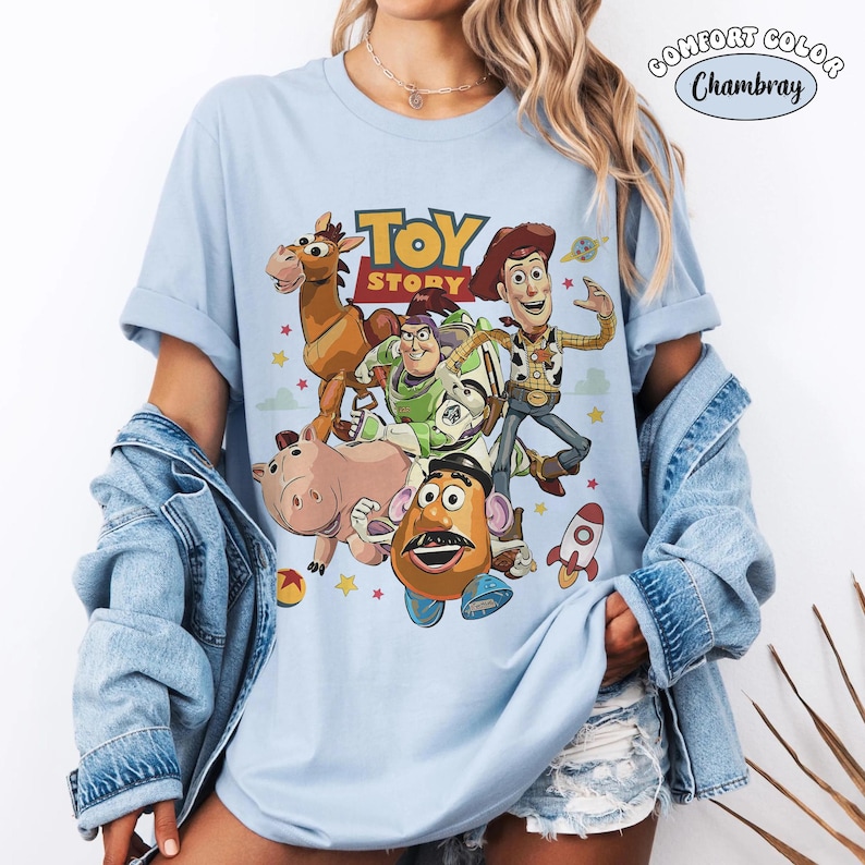 May include: Light blue Comfort Colors t-shirt featuring a graphic of characters from the animated film "Toy Story." The design includes Woody, Buzz Lightyear, Mr. Potato Head, and other characters. The words "Toy Story" are printed in yellow and red.
