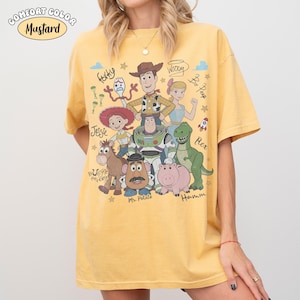 Może przedstawiać: Musztardowy t-shirt z nadrukiem graficznym przedstawiającym r&oacute;żne postacie animowane, w tym Chudego, Buzza Astrala, Jessie, Celka, Pana Bulwę, Hamm, Rexa, Pastereczkę i Sztućka, z ich imionami zapisanymi kursywą.