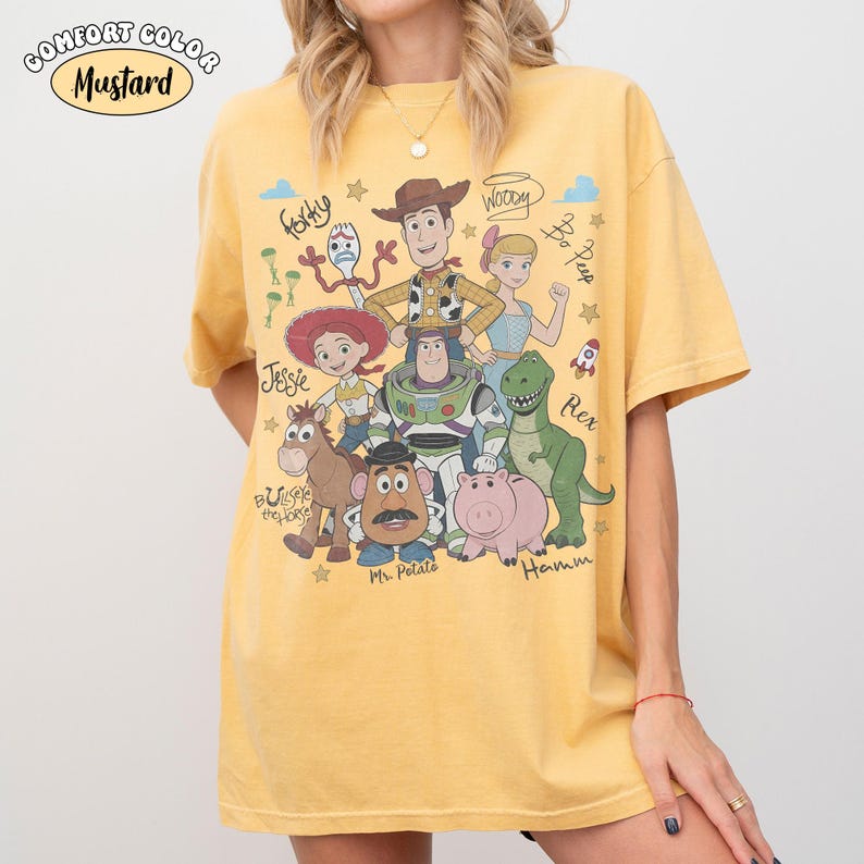 Może przedstawiać: Musztardowy t-shirt z nadrukiem graficznym przedstawiającym r&oacute;żne postacie animowane, w tym Chudego, Buzza Astrala, Jessie, Celka, Pana Bulwę, Hamm, Rexa, Pastereczkę i Sztućka, z ich imionami zapisanymi kursywą.
