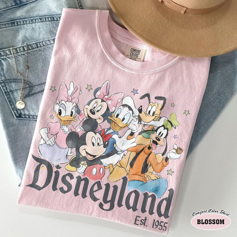 Pu&ograve; includere: Una t-shirt rosa chiaro con una stampa grafica di Topolino, Minnie, Paperino, Paperina, Pluto e Pippo. Il testo "Disneyland Est. 1955" &egrave; stampato in un carattere grigio scuro sotto i personaggi, circondato da piccole stelle colorate.