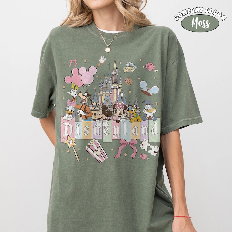 Peut inclure: T-shirt vert mousse Comfort Colors avec un motif Disneyland color&eacute;. Le motif comprend des personnages Disney, un ch&acirc;teau, des ballons et le logo Disneyland en couleurs pastel. Le t-shirt est de coupe d&eacute;contract&eacute;e.