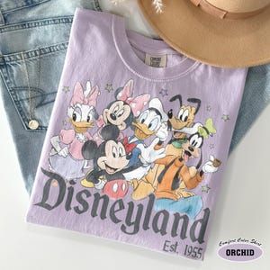 Pu&ograve; includere: Una t-shirt viola chiaro con una stampa grafica di Mickey Mouse, Minnie Mouse, Donald Duck, Daisy Duck, Goofy e Pluto. Il testo Disneyland Est. 1955 &egrave; stampato sotto i personaggi. La t-shirt presenta un'etichetta del marchio Comfort Colors.