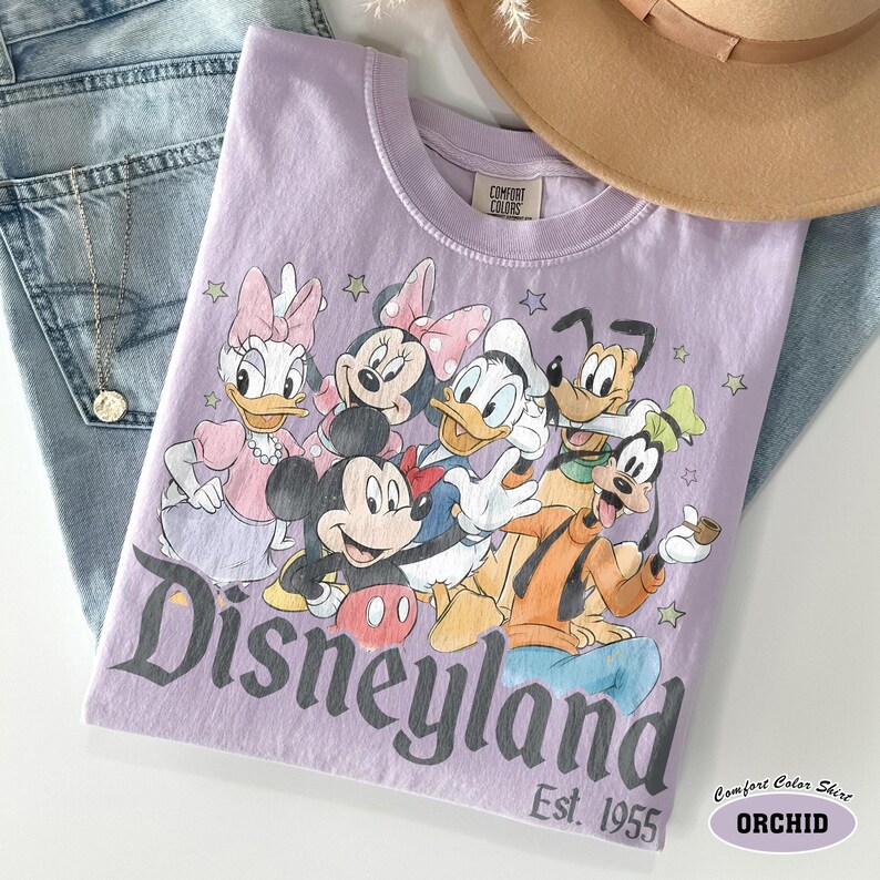 Pu&ograve; includere: Una t-shirt viola chiaro con una stampa grafica di Mickey Mouse, Minnie Mouse, Donald Duck, Daisy Duck, Goofy e Pluto. Il testo Disneyland Est. 1955 &egrave; stampato sotto i personaggi. La t-shirt presenta un'etichetta del marchio Comfort Colors.