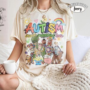 May include: An ivory t-shirt featuring a colourful graphic print with the word AUTISM in large, multi-coloured letters, followed by the text accept understand love. Below, a group of animated characters from Toy Story are illustrated.