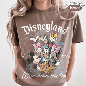 以下が含まれることがあります： エスプレッソブラウンのTシャツ。ミッキーマウス、ミニーマウス、ドナルドダック、デイジーダック、グーフィー、プルートのグラフィックがプリントされています。テキストは「Disneyland」「est. 1955」「Where dreams come true」と書かれています。右上にComfort Colorのラベルがあります。