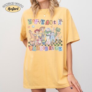 May include: Mustard-colored t-shirt featuring the phrase "You've Got a Friend in Me" with cartoon characters. The shirt has a crew neck and short sleeves. The text "Comfort Color" is in the upper left corner.