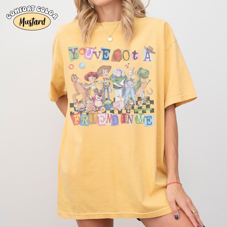 May include: Mustard-colored t-shirt featuring the phrase "You've Got a Friend in Me" with cartoon characters. The shirt has a crew neck and short sleeves. The text "Comfort Color" is in the upper left corner.