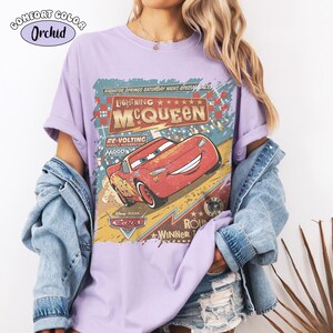 May include: A light purple t-shirt featuring a graphic of Lightning McQueen from the Disney Pixar Cars movie. The design includes the text "Lightning McQueen" and "Radiator Springs Saturday Night Special Race". The shirt is labeled "Comfort Color Orchid".
