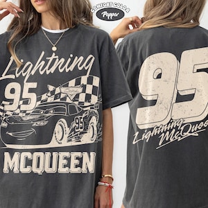May include: Dark gray t-shirt featuring a graphic of Lightning McQueen from the Cars movie. The front design includes the number 95, a checkered flag, and the name McQueen. The back of the shirt displays a large number 95 and the text "Lightning McQueen."