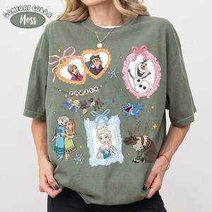 May include: Moss green Comfort Colors t-shirt featuring various characters and elements from the animated film "Frozen." The design includes framed portraits of characters, snowflakes, and the word "FROZEN" in a banner.