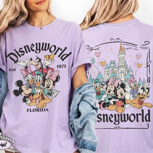 K&ouml;nnte beinhalten: Ein hellviolettes T-Shirt mit einem Grafikdruck von Disney-Figuren wie Mickey Maus, Minnie Maus, Donald Duck, Daisy Duck, Goofy und Pluto. Die Vorderseite zeigt den Text Disneyworld Est 1971 Florida. Die R&uuml;ckseite zeigt das Schloss.