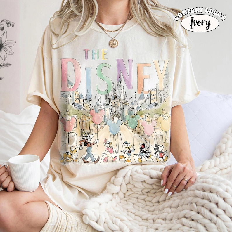 Pu&ograve; includere: T-shirt color avorio con una grafica colorata di personaggi Disney e la scritta "DISNEY". Il design include palloncini di Topolino e un castello. La maglietta &egrave; del marchio Comfort Color.