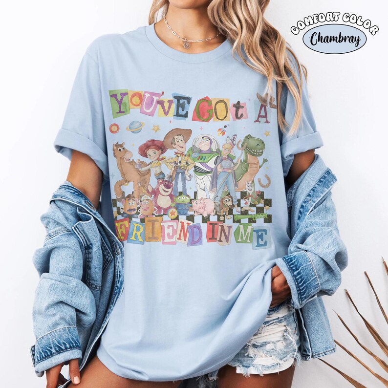 May include: Light blue Comfort Colors t-shirt featuring a colorful graphic of Toy Story characters. The text "YOU'VE GOT A FRIEND IN ME" is displayed above the characters in a playful font. The shirt has rolled-up sleeves and a relaxed fit.