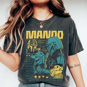 K&ouml;nnte beinhalten: Dunkelgraues T-Shirt mit einem gelben und t&uuml;rkisblauen Grafikdesign. Das Design zeigt den Text "MANDO" und Illustrationen des Mandalorianers und Grogu. Der Text "The Legend Continues" ist ebenfalls enthalten.