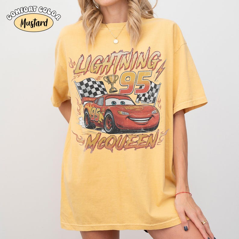 Puede incluir: Una camiseta de manga corta color amarillo mostaza con un gr&aacute;fico de un coche de carreras rojo con el n&uacute;mero 95, un trofeo y banderas a cuadros. El texto dice LIGHTNING 95 MCQUEEN. Una etiqueta en la esquina superior izquierda dice COMFORT COLOR MUSTARD.