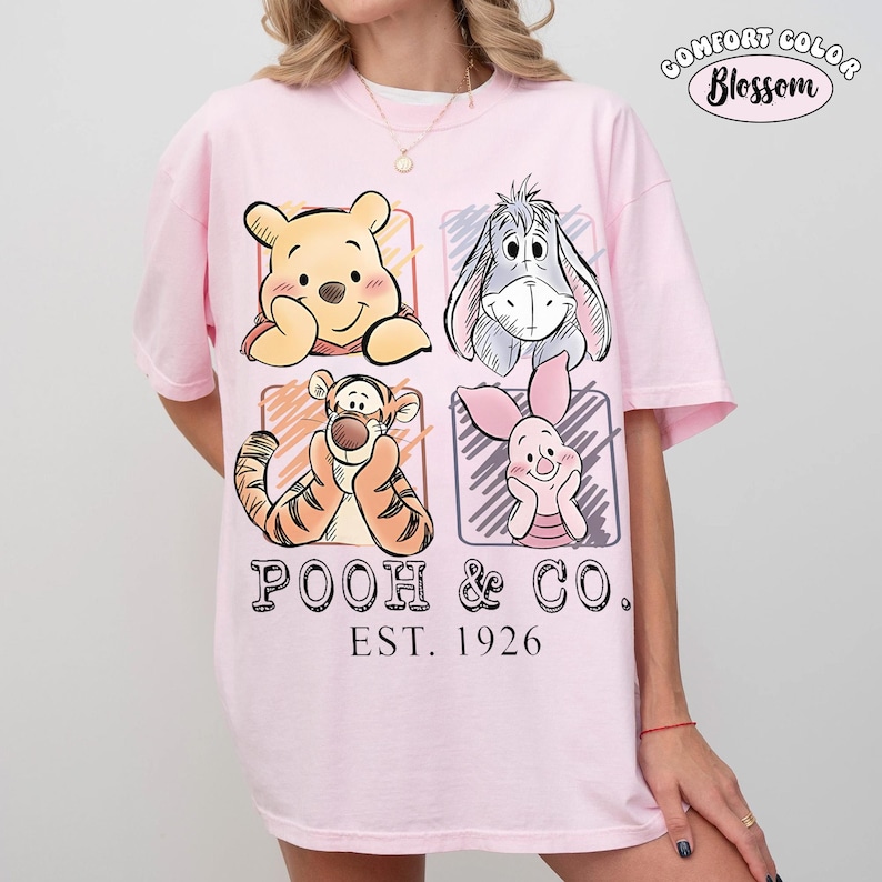 May include: Pink t-shirt featuring a graphic of Winnie the Pooh, Eeyore, Tigger, and Piglet in individual squares. The text "POOH & CO. EST. 1926" is printed below the characters. The shirt has short sleeves and a crew neck.