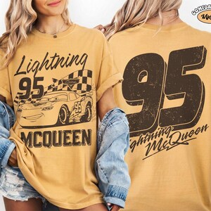 May include: Mustard-colored t-shirt featuring a graphic of Lightning McQueen from the movie Cars. The front design includes the text "Lightning 95 McQueen" and an image of the race car. The back of the shirt displays a large "95" and "Lightning McQueen".