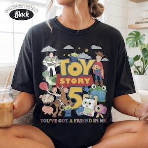 May include: A black crew neck t-shirt featuring a graphic print of Toy Story characters including Woody, Buzz Lightyear, and Jessie. The design includes the text 'TOY STORY 5' and 'YOU'VE GOT A FRIEND IN ME' in colorful lettering, with a 'COMFORT COLOR Black' label.
