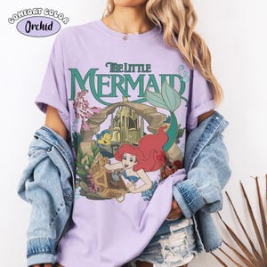 K&ouml;nnte beinhalten: Ein lila Comfort Colors T-Shirt mit einem Grafikdesign von Disneys Die kleine Meerjungfrau. Das Design zeigt Arielle, Fabius und Sebastian, mit dem Filmtitel in Gr&uuml;n. Das Shirt hat kurze &Auml;rmel und eine lockere Passform.