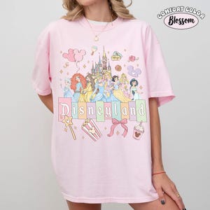 Puede incluir: Camiseta rosa Comfort Colors con un dise&ntilde;o de Disneyland. El dise&ntilde;o presenta princesas de Disney, un castillo y la palabra "Disneyland" en colores pastel. Otros elementos incluyen una varita m&aacute;gica, palomitas de ma&iacute;z y un lazo.