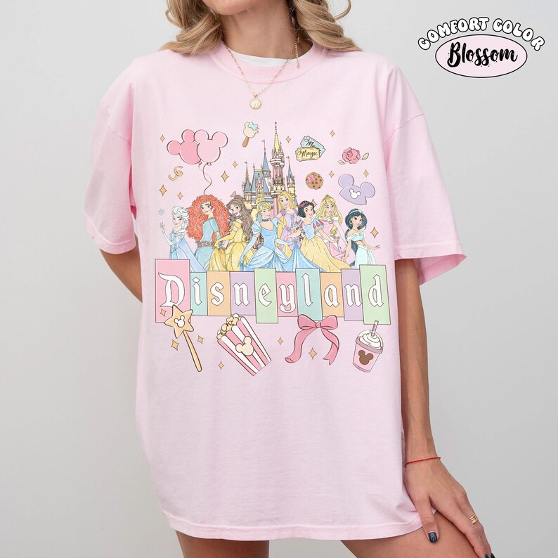 Puede incluir: Camiseta rosa Comfort Colors con un dise&ntilde;o de Disneyland. El dise&ntilde;o presenta princesas de Disney, un castillo y la palabra "Disneyland" en colores pastel. Otros elementos incluyen una varita m&aacute;gica, palomitas de ma&iacute;z y un lazo.