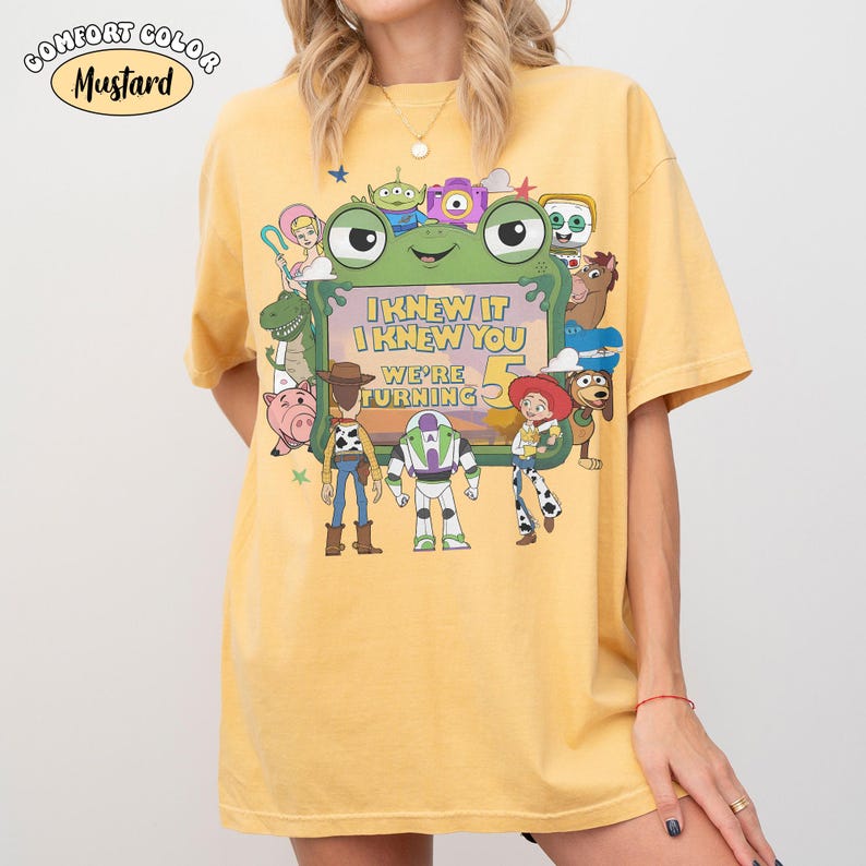 Puede incluir: Una camiseta de manga corta color amarillo mostaza con un estampado gr&aacute;fico de varios personajes animados rodeando un marco verde con forma de rana. El texto dentro del marco dice, I KNEW IT I KNEW YOU WE'RE TURNING 5. Cuello redondo.