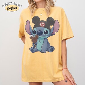 May include: A mustard yellow short-sleeve t-shirt featuring a graphic of the blue character Stitch wearing a black hat with mouse ears and holding a chocolate-covered treat. The text COMFORT COLOR MUSTARD is printed in the upper left corner.