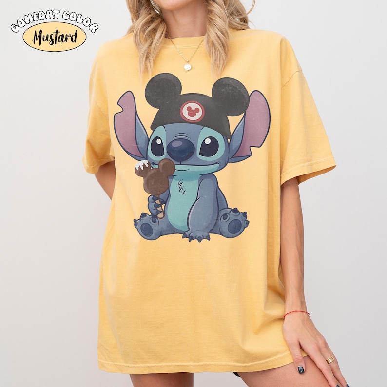 May include: A mustard yellow short-sleeve t-shirt featuring a graphic of the blue character Stitch wearing a black hat with mouse ears and holding a chocolate-covered treat. The text COMFORT COLOR MUSTARD is printed in the upper left corner.