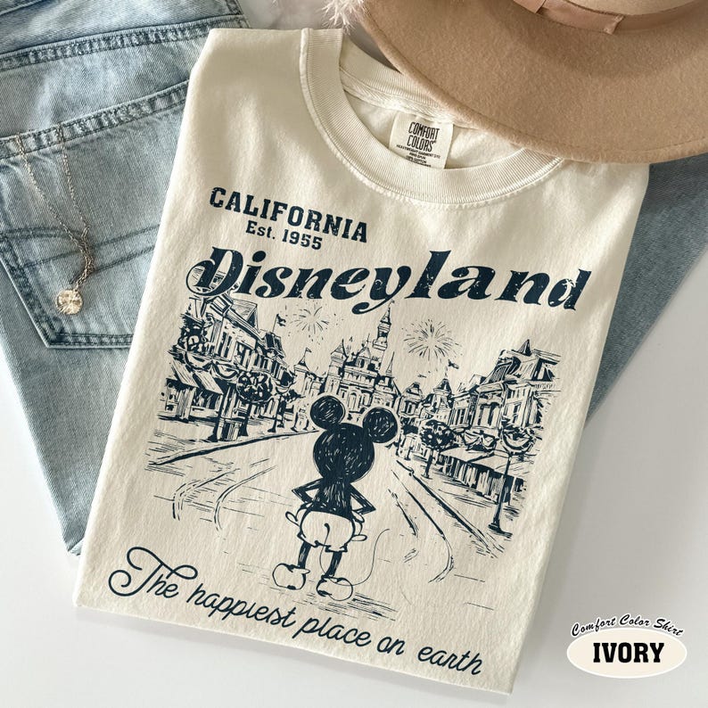 May include: Ivory-colored Comfort Colors t-shirt featuring a Disneyland graphic. The design includes the text "CALIFORNIA Est. 1955 Disneyland" and "The happiest place on earth" with a Mickey Mouse illustration. The shirt is laid on top of blue jeans.