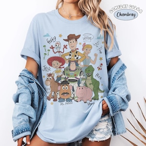 Może przedstawiać: Jasnoniebieski t-shirt z nadrukiem graficznym przedstawiającym postacie z Toy Story, w tym Chudego, Buzza Astrala, Jessie, Rexa, Hamm, Pana Bulwę, Pastereczkę i Sztućka, z ich imionami zapisanymi kursywą. Widoczny jest tekst Comfort Color Chambray.