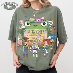 Puede incluir: Una camiseta de color verde musgo con un gran gr&aacute;fico de varios personajes animados que rodean un marco central con forma de rana. El texto dentro del marco dice, I KNEW IT I KNEW YOU WE'RE TURNING 5. El dise&ntilde;o incluye figuras como un vaquero, un guardi&aacute;n espacial y una vaquera.