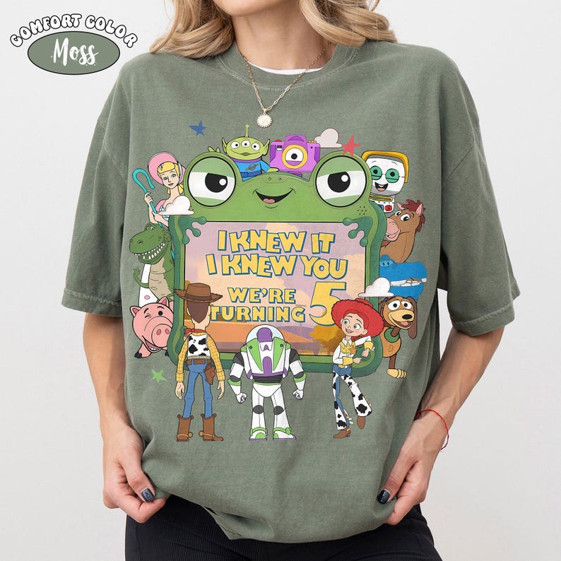 Puede incluir: Una camiseta de color verde musgo con un gran gr&aacute;fico de varios personajes animados que rodean un marco central con forma de rana. El texto dentro del marco dice, I KNEW IT I KNEW YOU WE'RE TURNING 5. El dise&ntilde;o incluye figuras como un vaquero, un guardi&aacute;n espacial y una vaquera.