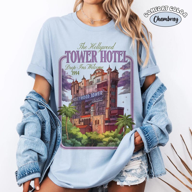 Op de afbeelding: Lichtblauw Comfort Colors Chambray T-shirt met een afbeelding van het Hollywood Tower Hotel. Het ontwerp bevat de tekst "The Hollywood Tower Hotel" en "Drop-Ins Welcome 1994". Het shirt heeft opgerolde mouwen.