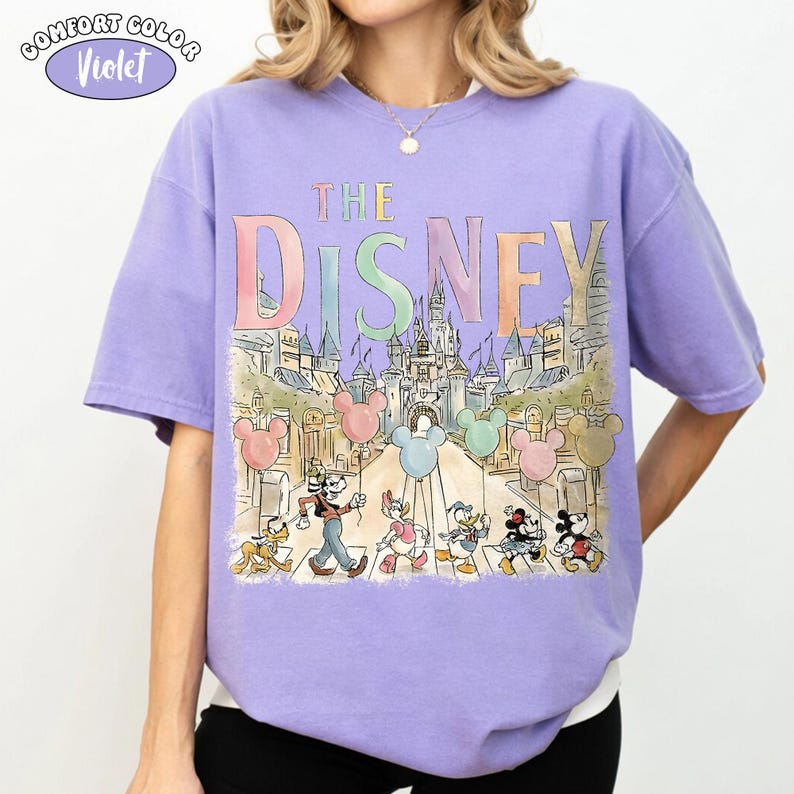 Pu&ograve; includere: Una maglietta Comfort Colors viola con la scritta "DISNEY" in colori pastello sopra un'illustrazione di cartoni animati di personaggi Disney che attraversano una strada di fronte a un castello. Topolino, Paperino e altri personaggi sono raffigurati.