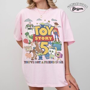 May include: A light pink t-shirt featuring a large graphic print of Toy Story characters including Woody, Buzz Lightyear, and Rex. The text reads TOY STORY 5 and YOU'VE GOT A FRIEND IN ME. A small logo in the top right corner says COMFORT COLOR Blossom.