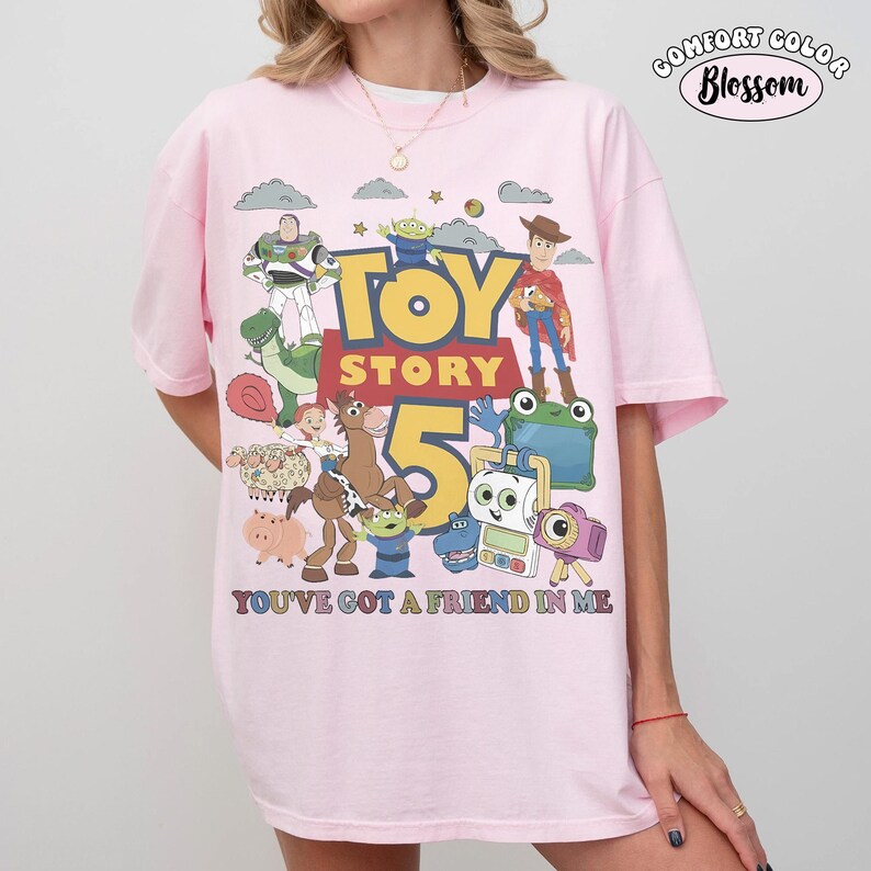 May include: A light pink t-shirt featuring a large graphic print of Toy Story characters including Woody, Buzz Lightyear, and Rex. The text reads TOY STORY 5 and YOU'VE GOT A FRIEND IN ME. A small logo in the top right corner says COMFORT COLOR Blossom.