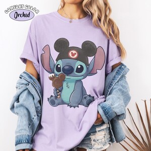 May include: A light purple t-shirt featuring a graphic of the character Stitch wearing a black Mickey Mouse ear hat and holding a chocolate Mickey-shaped ice cream bar. The text Comfort Color Orchid is printed in the upper left corner of the image.
