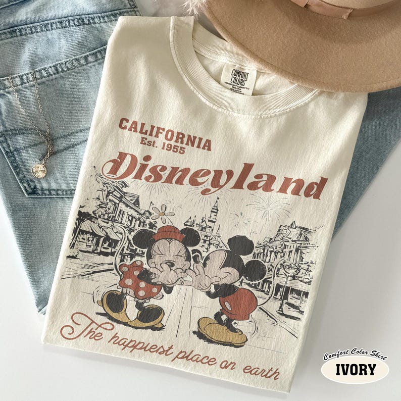 May include: An ivory t-shirt featuring a graphic print of Mickey and Minnie Mouse in front of a castle. The text reads California Est. 1955 Disneyland and The happiest place on earth. The shirt has a Comfort Colors label at the inner neckline.