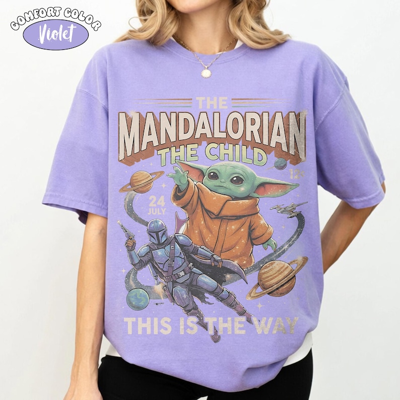 May include: A violet short-sleeve t-shirt featuring a graphic print of The Mandalorian and The Child in space. The text reads THE MANDALORIAN, THE CHILD, 24 JULY, and THIS IS THE WAY. A small circular label in the corner says COMFORT COLOR Violet.