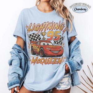 Puede incluir: Una camiseta azul claro con un gr&aacute;fico del coche de carreras rojo Lightning McQueen, el n&uacute;mero 95, un trofeo dorado y banderas a cuadros. El texto dice LIGHTNING MCQUEEN y 95. Una etiqueta circular en la esquina dice COMFORT COLOR Chambray.