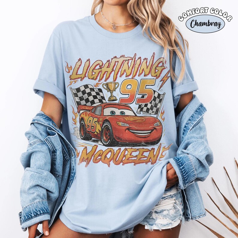 Puede incluir: Una camiseta azul claro con un gr&aacute;fico del coche de carreras rojo Lightning McQueen, el n&uacute;mero 95, un trofeo dorado y banderas a cuadros. El texto dice LIGHTNING MCQUEEN y 95. Una etiqueta circular en la esquina dice COMFORT COLOR Chambray.