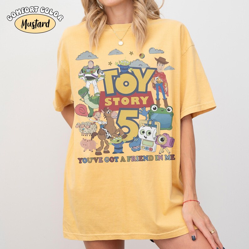 May include: A mustard yellow short-sleeve t-shirt featuring a graphic print of Toy Story characters including Woody, Buzz Lightyear, and Rex. The text reads "TOY STORY 5" and "YOU'VE GOT A FRIEND IN ME". A label in the corner says "COMFORT COLOR MUSTARD".