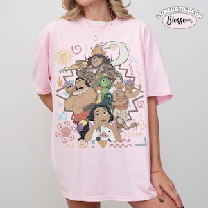 Puede incluir: Una camiseta rosa claro con un estampado gr&aacute;fico de personajes de la pel&iacute;cula Moana, incluyendo a Moana, Maui, Pua, Heihei y Gramma Tala. El dise&ntilde;o incluye patrones de inspiraci&oacute;n tribal, s&iacute;mbolos y el texto Comfort Color Blossom.