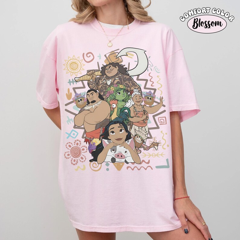 Puede incluir: Una camiseta rosa claro con un estampado gr&aacute;fico de personajes de la pel&iacute;cula Moana, incluyendo a Moana, Maui, Pua, Heihei y Gramma Tala. El dise&ntilde;o incluye patrones de inspiraci&oacute;n tribal, s&iacute;mbolos y el texto Comfort Color Blossom.