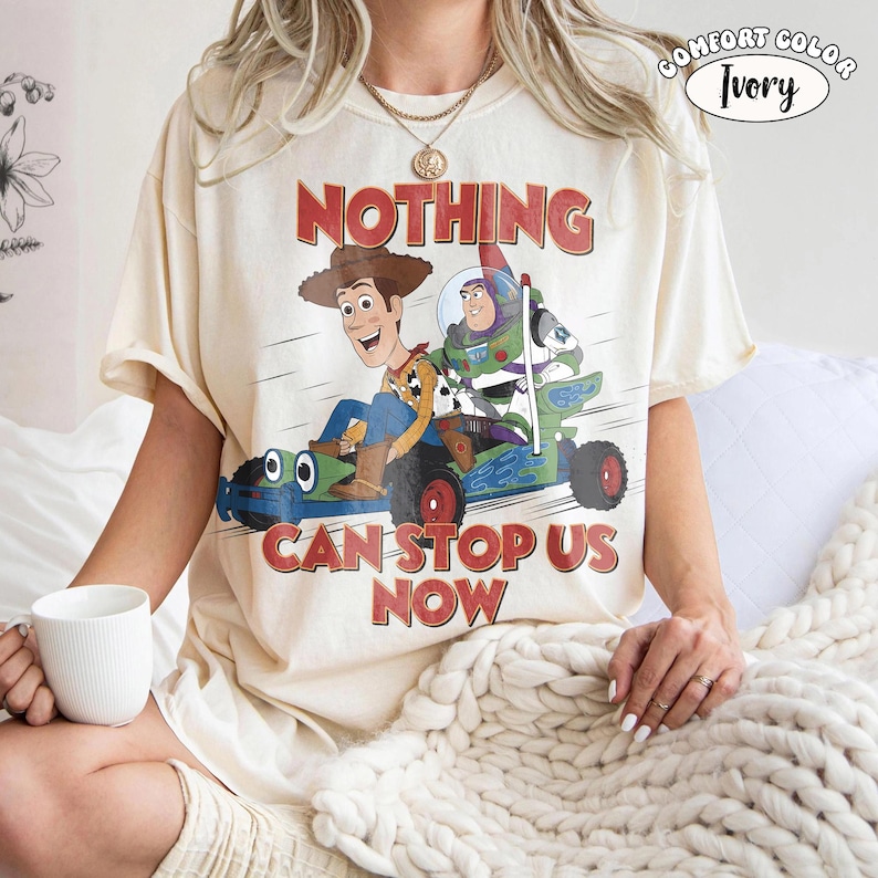 May include: Ivory-colored t-shirt featuring a graphic of Woody and Buzz Lightyear in a race car, with the text "NOTHING CAN STOP US NOW" in red. The shirt is a Comfort Colors brand.