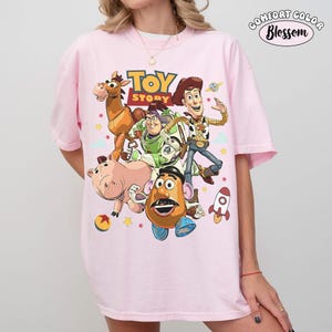 May include: A light pink Comfort Colors t-shirt featuring a colorful graphic of Toy Story characters, including Woody, Buzz Lightyear, and Mr. Potato Head. The words "TOY STORY" are printed above the characters. The shirt has short sleeves.